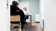 En kvinna sitter bakom ett skrivbord och talar med en ung svartklädd man.