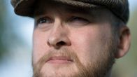 Henkilö katsoo eteenpäin ulkona, yllään ruskea lippalakki ja ruudullinen kauluspaita. Tausta on epätarkka.