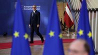 A man in a dark suit seen in semi-profile walking in front of a sign saying European Council, with blurry blue EU flags in the foreground.