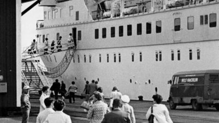 Mustavalkoisessa kuvassa ihmiset kävelevät matkalaukkuineen isoon matkustajalaivaan.