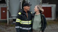 Två vuxna personer står utomhus framför två stora silos och en byggnad med röda dörrar.