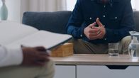 Man sitting in sofa facing another man with a notebook in his lap. A coffee table with a glass of water is seen between them.