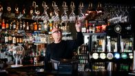 A bald middle-aged man stands behind a bar, reaching up for a glass with many bottles behind him.