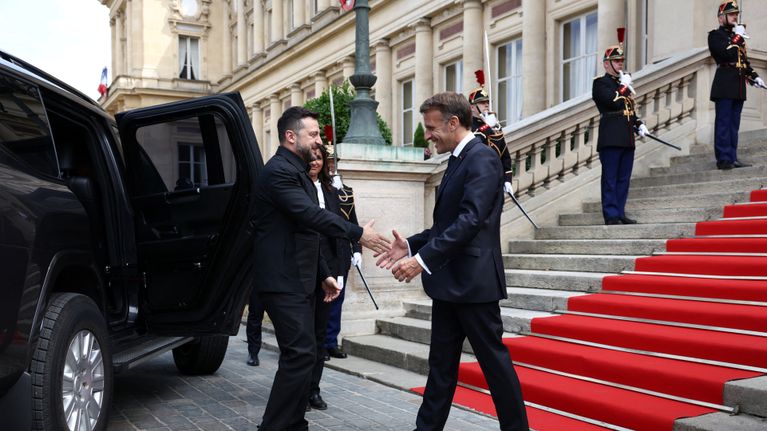 Volodymyr Zelenskyi ja Emmanuel Macron kättelevät portaiden edessä, Zelenskyin takana auto, jonka ovi on auki.