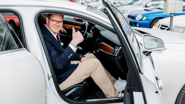 Mies istuu valkoisen auton etupenkillä parkkipaikalla ja näyttää peukaloa.