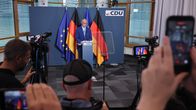 En man i blå kostym ser liten ut vid ett talarpodium framför Tysklands och EU:s flaggor. På talarpodiet och på väggen bakom står det CDU.