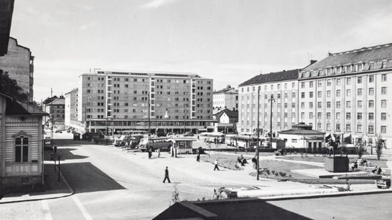 Ett torg med bussar, bilar och människor omges av flerbostadshus och äldre trähus.