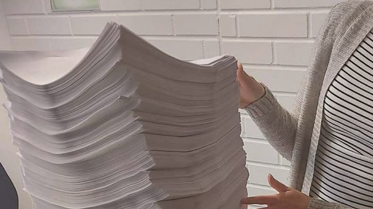 Kuvassa näkyy suuri pino paperiarkkeja, jotka ovat kaareutuneet hieman painon alla. Henkilö, jonka kasvot eivät näy, pitelee pinoa varoen molemmin käsin. Taustalla näkyy valkoinen tiiliseinä ja vasemmassa alakulmassa pieni kasvi. Henkilöllä on yllään raidallinen paita ja vaalea neuletakki. Kuvan alareunassa näkyy lisäksi muovilaatikoita mahdollisesti toimistotarvikkeita varten.