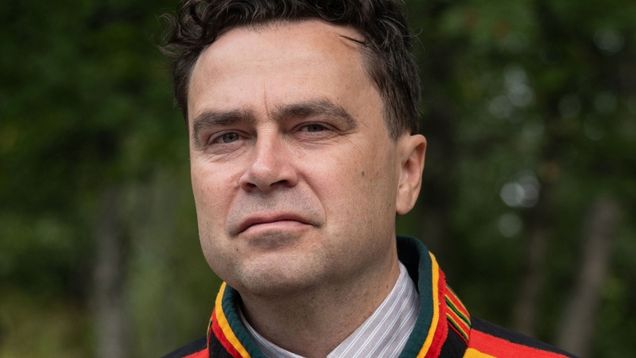 En man med mörkt lockigt hår och traditionell samisk kolt står utomhus med träd i bakgrunden. 
