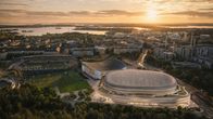 Ilmakuva suuresta stadionista ja sitä ympäröivistä rakennuksista ilta-auringon aikaan; taustalla merimaisema.