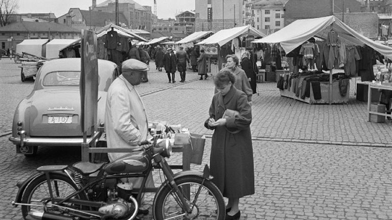 Svartvit bild. Två personer står vid en motorcykel på ett torg med marknadsstånd, bilar och byggkran i bakgrunden.