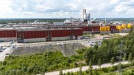Skogsindustriområde med fabriksbyggnader och kraftöverföringsledningar, beläget i en grönskande miljö under en molnig himmel.