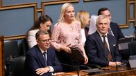 Riikka Purra i en blommig skjorta står och talar i riksdagen. Bredvid henne sitter kostymklädda Petteri Orpo och Anders Adlercreutz.