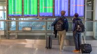 Two people with small suitcases looking at a large flight schedule screen at an airport.