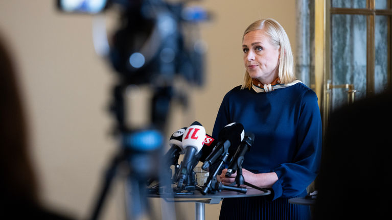 A blonde woman, Elina Valtonen, stands in front of microphones.