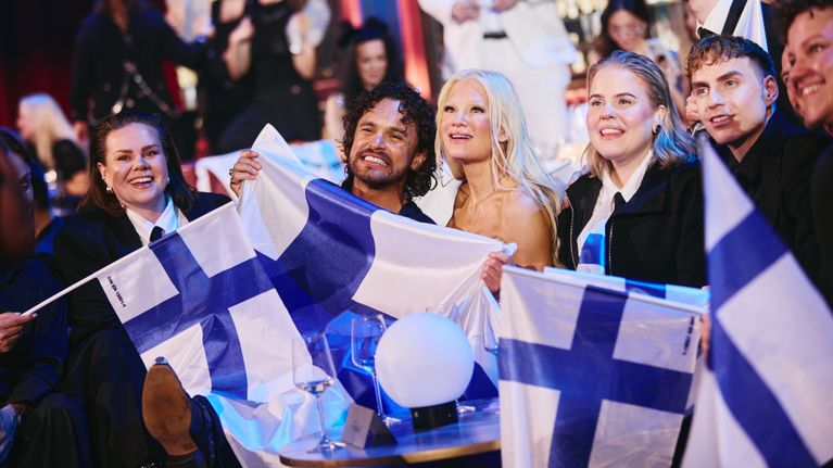 En grupp människor sitter tillsammans inomhus och håller upp finska flaggor.
