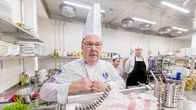 En kock i vit kockrock och hög vit kockhatt står i ett storkök. Två andra personer i kockkläder syns i bakgrunden.