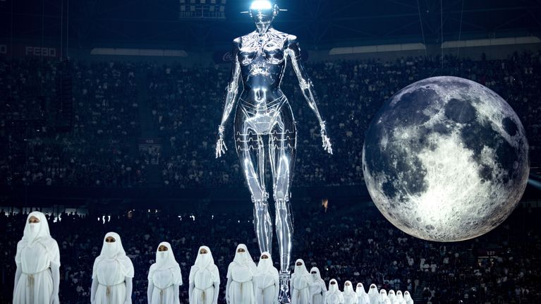 Kuva The Weekndin keikalta Amsterdamista, jossa lavalla marssi valkopukuisia ihmisiä robottipatsaan ja kuun alla.