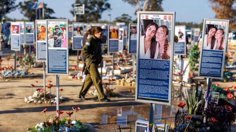 Skyltar på personer som dött står på en jordplan. Vid skyltarna finns blommor. En israelisk soldat går bland skyltarna.
