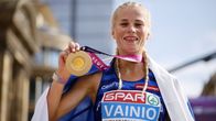 Alisia Ainio håller i en guldmedalj och ler medan hon tittar in i kameran. Hon har Finlands flagga över axlarna.