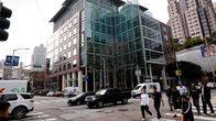 Useita ihmisiä ylittää suojatietä vilkkaassa risteyksessä, taustalla lasiseinäinen toimistorakennus ja pilvenpiirtäjiä.