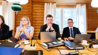 Three people sit at at table in a log cabin behind laptops and folders. 