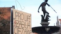 Käsi pitelee pahvikylttiä, jossa lukee "PRO LIFE ZABIJA CIEZARNE", taustalla suihkulähde ja patsas.