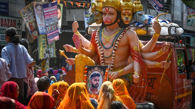 Värikkäästi pukeutuneet ihmiset pitelevät miehen valokuvaa kadulla suuren hindulaisen jumalhahmon patsaan ympärillä