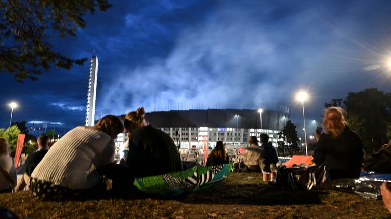 Ihmisiä istuu nurmikolla iltahämärässä Helsingin Olympiastadionin ulkopuolella, taustalla näkyy valaistu stadion ja savua yläpuolella Coldplayn keikan aikana 31. heinäkuuta 2024.