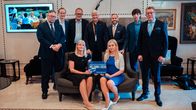 Nine people posing for a photo; two people sat on a sofa and seven people standing behind them. They are holding a sign which reads "ROAD TO HELSINKI 2030".