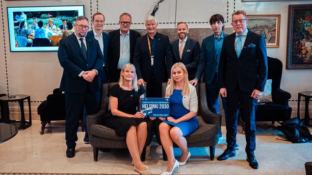 Nine people posing for a photo; two people sat on a sofa and seven people standing behind them. They are holding a sign which reads "ROAD TO HELSINKI 2030".