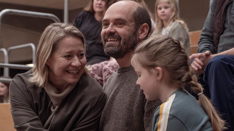 En familj med mamma, pappa och dotter sitter leende tätt tillsammans på en läktare i en sporthall.