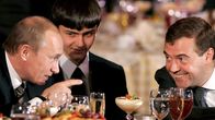 Vladimir Putin osoittaa hymyilevää Dmitri Medvedevia päivällispöydän lasien yli.