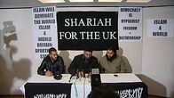 Islam4UK-järjestön johtaja istuu kahden jäsensä kanssa pitämässä lehdistötilaisuutta. Taustalla paperiarkkeja ja julisteita, joissa lukee muun muassa "Shariah for the UK" ja "Islam will Dominate the World".