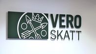 Vero- ja skatt-tekstit logosymbolin kanssa valkoisella seinällä. Tekstit ovat hieman irti seinästä, jolloin niiden alle muodostuu pienoinen varjo.