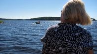 Nainen (selin) istuu järven rannalla, katselee järvellä olevaa soutuvenettä.