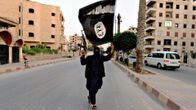 Mustiin pukeutunut aseistaunut henkilö pitelee ISIS-lippua ilmassa.
