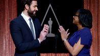 John Krasinski ja Cheryl Boone Isaacs katsovat toisiaan ja taputtavat käsiään teatterilavalla. Taustalla elokuva-akatemian logo.