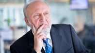 Hans-Werner Sinn katsoo kameran ohi mietteliäänä, pitäen oikean kätensä sormia oikeanpuoleisella poskellaan ja ja ylähuulellaan. Hänellä on tumma puku liivien kera, valkoinen kauluspaita ja kirkkaansininen kravatti. Sinnin parta on valkoinen ja hiusraja on korkealla otsalla.