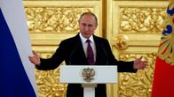 Putin puhuu korokkeen takaa ja levittää juuri käsiään. Putinilla on tumma asu ja viininpunainen kravatti. Taustalla näkyy suuri kullattu ja koristeltu ovi ja kuvan oikeassa ja vasemmassa reunassa Venäjän liput. Korokkeessa on Venäjän vaakunaeläin, kaksipäinen kotka.