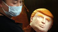 Mies hymyilee, katsoo alaviistoon hengityssuojain suun edessä, kädessään Trumpin kasvoja esittävä kuminaamari. 
