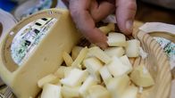 Miehen käsi poimii juustonpalaa vadilta, johon on kuutioitu puolikas juustokiekosta. 