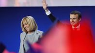 Brigitte Trogneux ja Emmanuel Macron seisovat esiintymislavalla. Macron heiluttaa kättään. Etualalla joku heiluttaa jonkinlaista punaista vaatetta, joka näkyy kuvassa punaisen värin leiskahduksena.