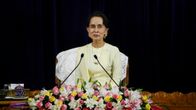 Aung San Suu Kyi istuu kukkalaitteen ja kahden mikrofonin takana vaaleankeltaisessa asussa tummaa taustaa vasten.