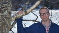 Mies pitää koivun oksasta kiinni rantamaisemassa ja katsoo suoraan kameraan