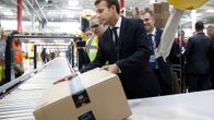 Emmanuel Macron nostaa Amazonin paketin liukuhihnalle.