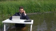 Mies puhuu puhelimeen työpöydän ääressä rantavedessä, kaislikon edessä,