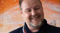 Jari Peltomäki lukee Seiskaa oranssin taustan edessä. 