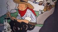 Piirros, joss cowboy-asuinen Tintti istuu kivellä Milou-koira rinnallaan ja heidän takanaan hiipii kirveillä aseistautuneita intiaaneja.