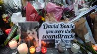 Keko muovi- ja paperikääröissä olevia kukkia ja niiden edessä kyntilöitä. Bowien valokuvan vieressä iso kortti, jossa lukee englanniksi: "David Bowie, olit elämämme elokuvan ääniraita".
